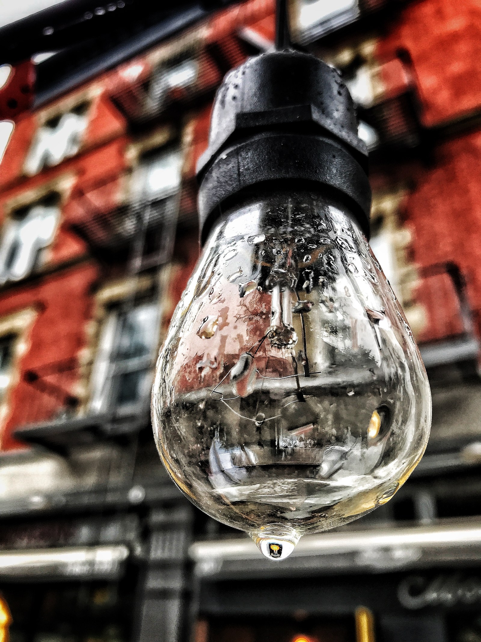 A rain-covered light bulb photographed close, red brick and fire escapes soft behind it.