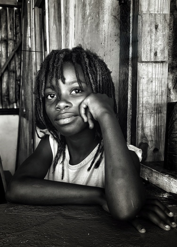 A young girl sitting with her head resting against her hand, looking outward, in a wooden shelter.