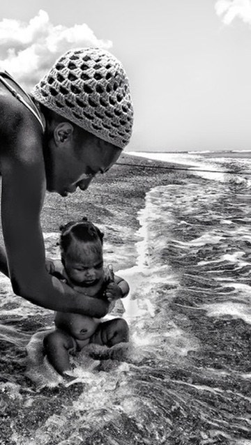 A woman steadying a baby girl as the sea washes around them at the shoreline.