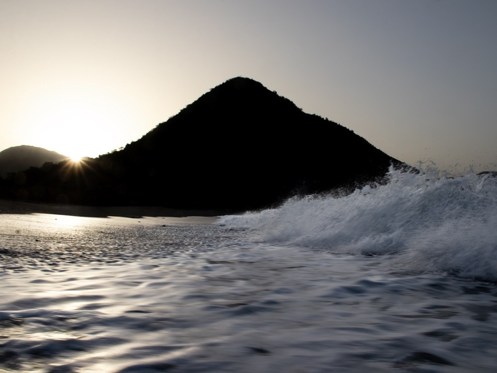 A dark mountain silhouetted against a low sun, with a wave breaking across the foreground.