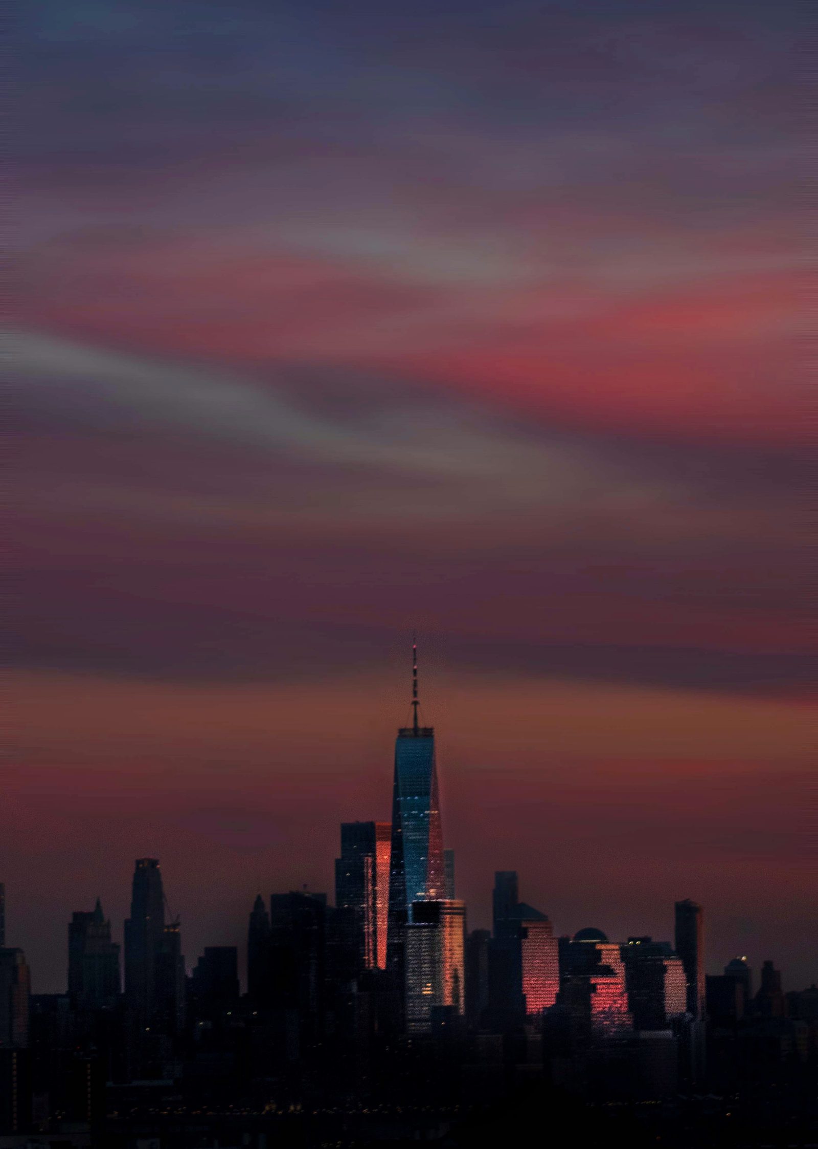 A city skyline at dusk seen from far away, a single spired tower rising under long bands of pink and violet cloud.