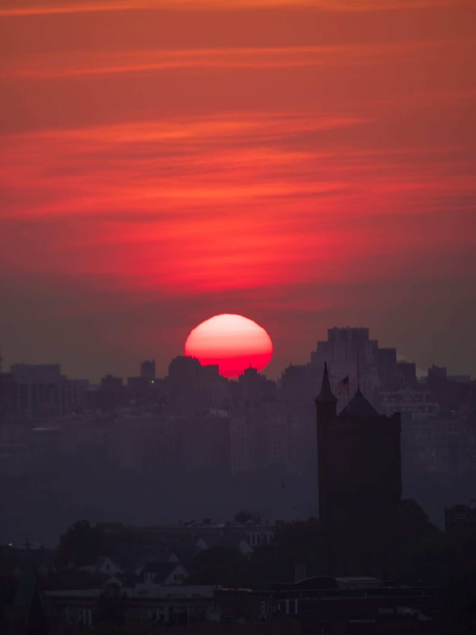 A red sun sinking behind distant apartment towers in haze, the dark silhouette of a turreted building in the foreground.