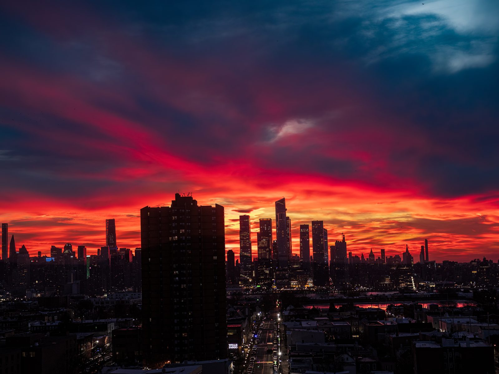 The skyline in silhouette under a burning red and deep blue sky, a tall dark apartment block in front and a lit street running toward the river.