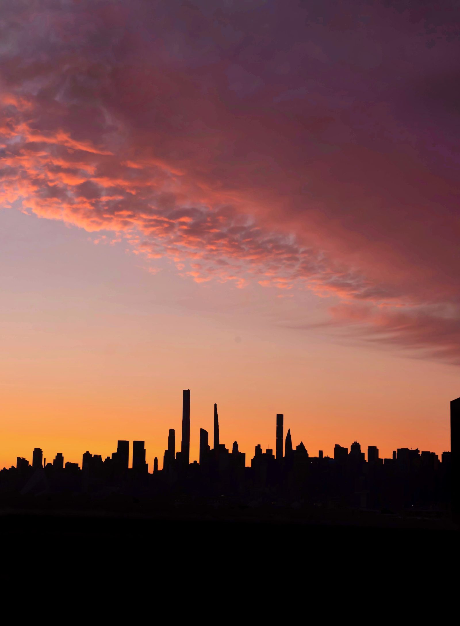A pink and violet cloud bank over a pale orange sky, the skyline a flat black silhouette along the bottom of the frame.
