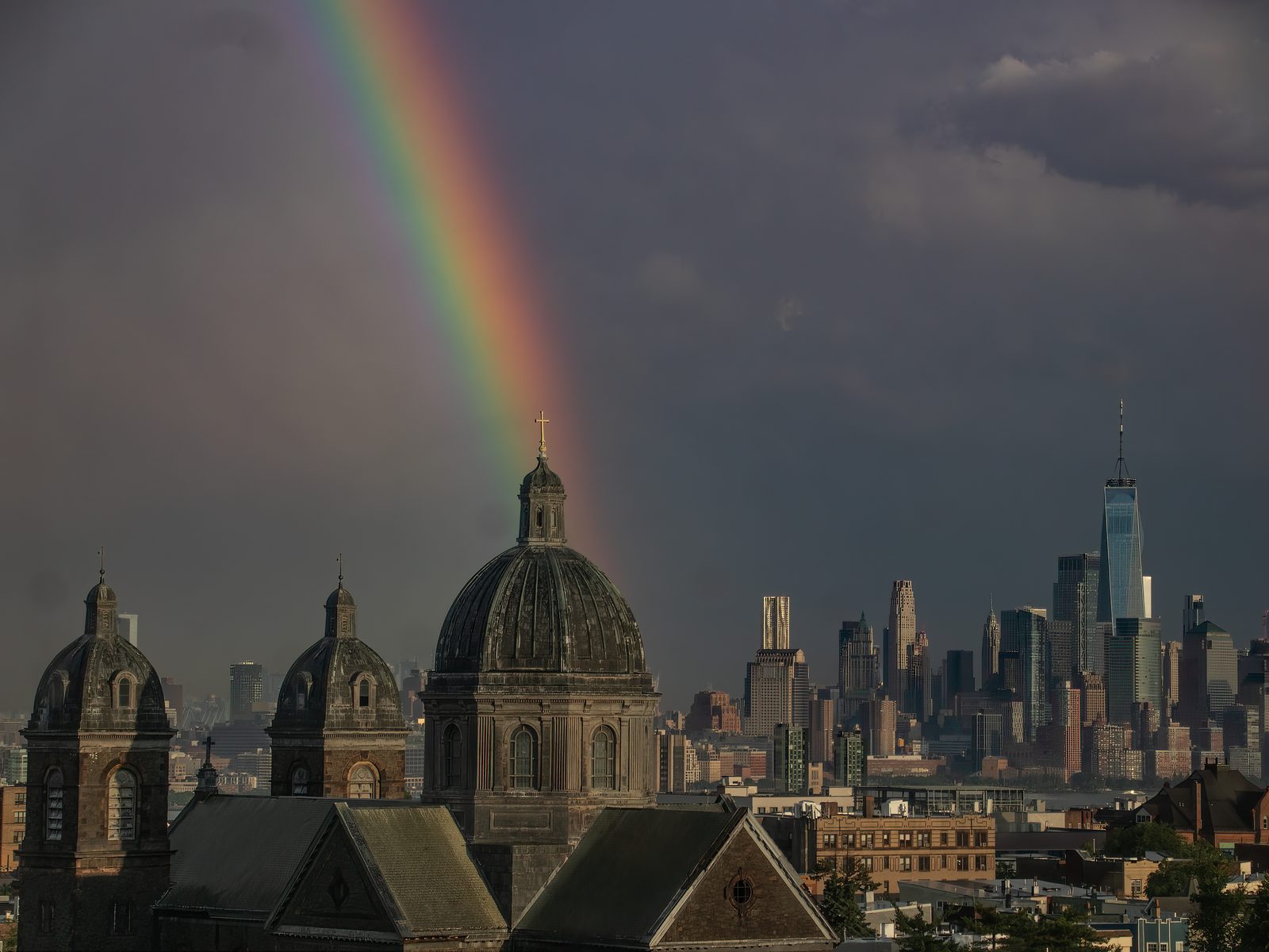 A rainbow coming down through a dark grey sky onto the dome and cross of a church, with the lit towers of the city beyond.