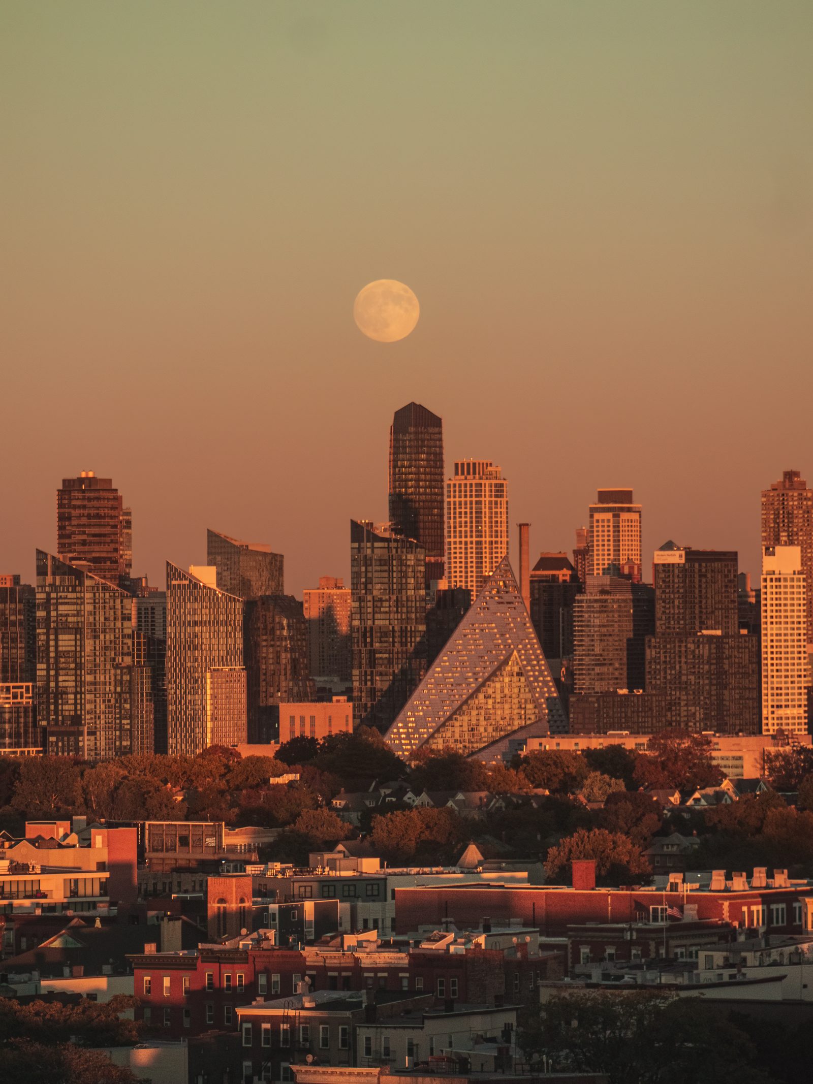 A full moon over a cluster of glass towers and a pyramid-shaped building, rooftops glowing in evening light below.
