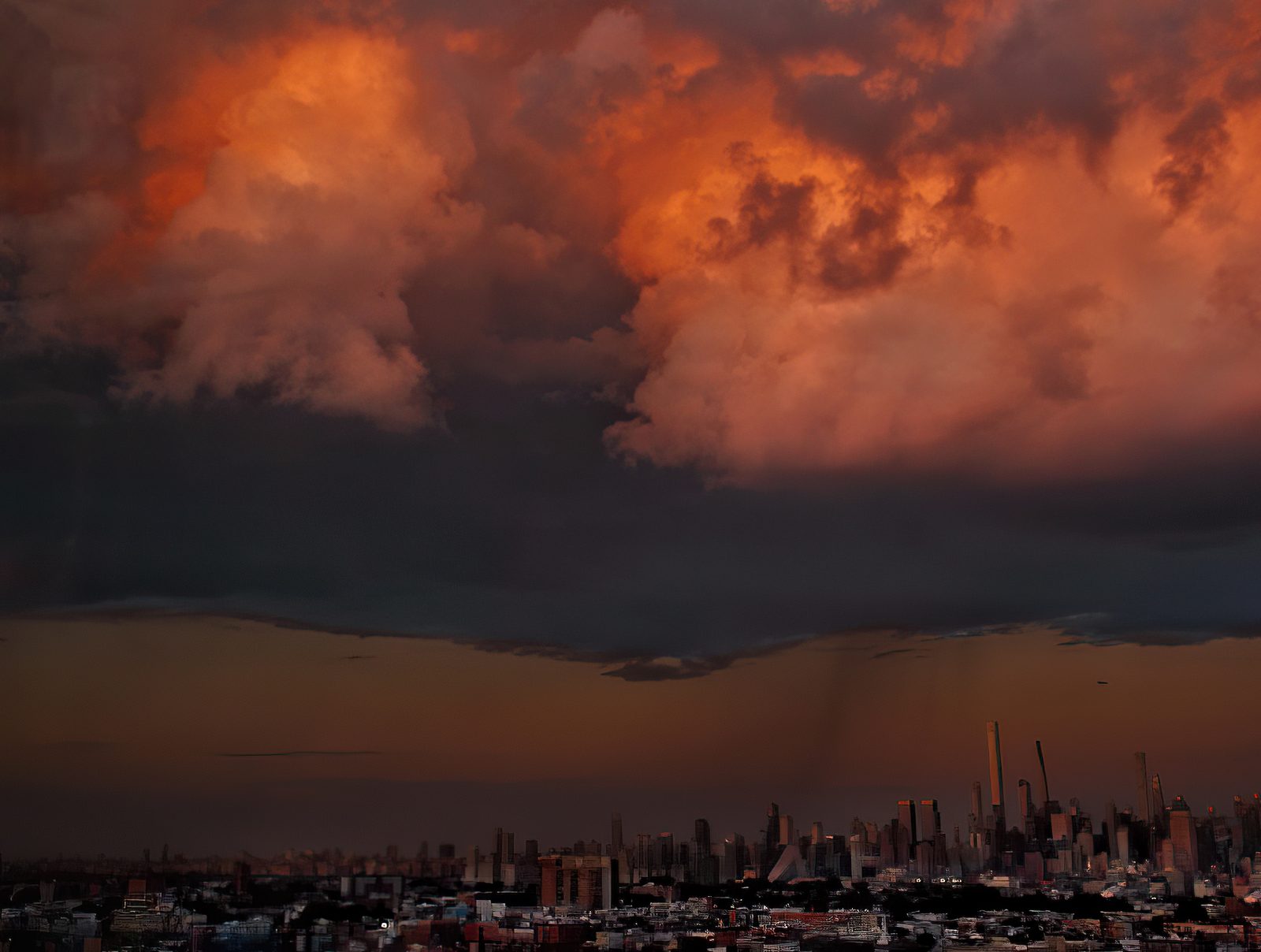 Towering orange storm clouds over a dark sky, the city small along the bottom edge with a thin shaft of rain.
